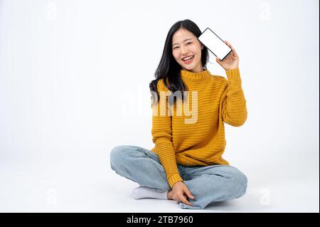 Belle femme asiatique dans des vêtements décontractés assis sur le sol sur un fond blanc isolé et montre son smartphone maquette d'écran blanc à la caméra. Banque D'Images