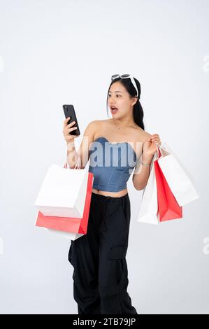 Une femme asiatique élégante porte ses sacs à provisions et regarde l'écran de son smartphone avec un visage magnifique, surpris par la promotion en ligne disque Banque D'Images