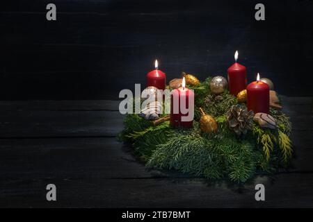 Lumière dans l'obscurité sur le quatrième avènement, couronne verte naturelle avec des bougies rouges, quatre brûlent, décoration de Noël et biscuits, fond en bois sombre, Banque D'Images