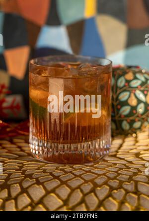 Tequila Negroni en verre à boule basse avec de la glace et de la chaux devant les décorations de Noël, y compris les boules et le ruban rouge sur le tapis doré Banque D'Images