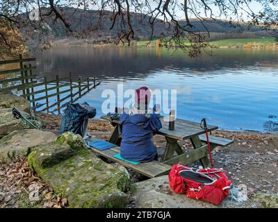 Femme assise à la table de pique-nique en bois sur les rives du lac Ullswater dans le English Lake District vu de Sandwick en automne, Cumbria, Angleterre, Royaume-Uni Banque D'Images