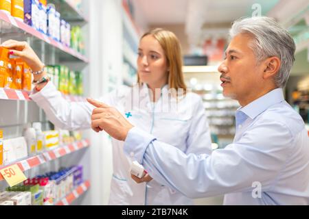 Jeune pharmacienne en uniforme blanc aidant le client masculin senior à acheter et choisir des médicaments sur les étagères du magasin Banque D'Images