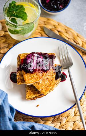 Pile de crêpes maison garnie de confiture sucrée de myrtilles et une cuillerée de crème sure et de boisson à l'eau de citron servie sur une assiette blanche avec un bord bleu. Banque D'Images