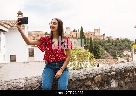 Jeune femme souriante habillée en casuals prenant selfie avec smartphone tout en étant assise sur le mur de soutènement avec Alcazaba forteresse en arrière-plan à Grenade, Banque D'Images