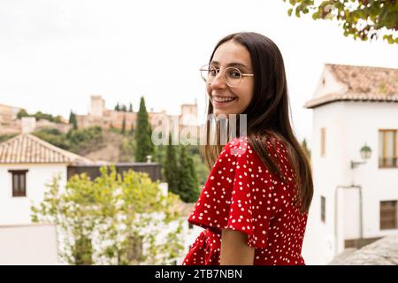 Vue latérale de la jeune femme touriste heureuse debout tout en regardant la caméra avec la forteresse Alcazaba en arrière-plan à Grenade, Espagne Banque D'Images