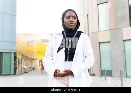 Femme entrepreneure afro-américaine musulmane sérieuse vêtue de hijab et de formels tenant un ordinateur portable et regardant loin tout en se tenant dans la rue Banque D'Images