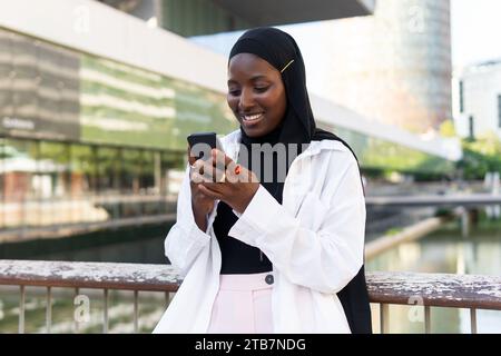 Joyeuse jeune femme d'affaires afro-américaine dans la navigation hijab sur téléphone intelligent tout en se tenant contre le bâtiment moderne Banque D'Images