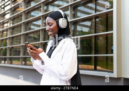 Joyeuse jeune femme d'affaires afro-américaine en foulard noir écoutant de la musique sur des écouteurs et utilisant smartphone contre la construction Banque D'Images