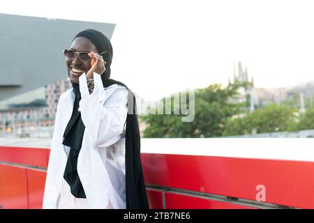 Élégant jeune femme d'affaires musulmane joyeuse en foulard noir traditionnel portant des lunettes de soleil tout en se tenant par le mur de soutènement contre le ciel Banque D'Images