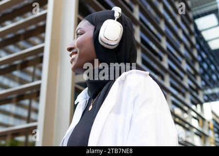 Vue de côté de souriante musulmane jeune femme d'affaires afro-américaine dans le hijab écoutant de la musique sur des écouteurs et regardant loin contre le bâtiment Banque D'Images