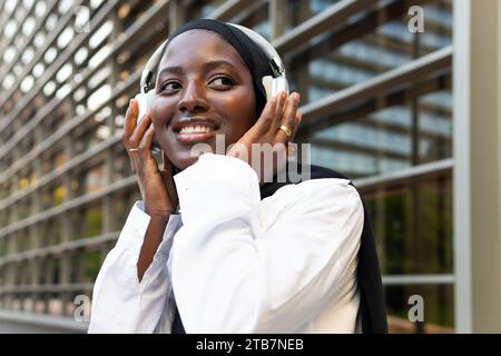 Gros plan de souriante musulmane jeune femme d'affaires afro-américaine dans le hijab écoutant de la musique sur des écouteurs et regardant loin contre la construction Banque D'Images
