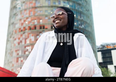 Vue à angle bas de l'entrepreneur féminin afro-américain à la mode en hijab noir et lunettes de soleil assis contre le bâtiment moderne dans la ville Banque D'Images