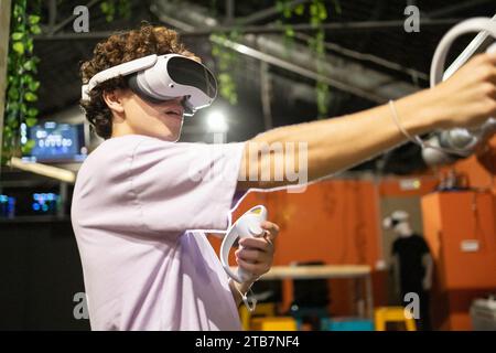 Un jeune joueur enthousiaste est entièrement immergé dans un jeu de réalité virtuelle, portant un casque et utilisant des contrôleurs de mouvements. Banque D'Images