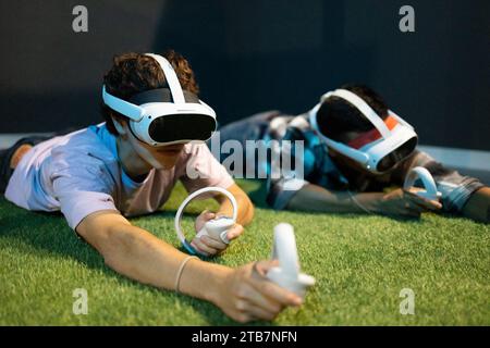 Un jeune homme est allongé sur du gazon artificiel, engagé dans des jeux de réalité virtuelle avec un casque VR et des manettes portables Banque D'Images