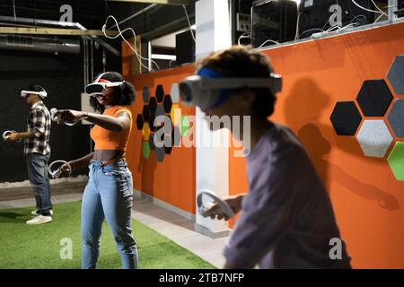 Vue latérale de jeunes amis multiraciaux engagés dans des jeux de réalité virtuelle portant des casques VR et utilisant des manettes portables dans un cadre intérieur Banque D'Images