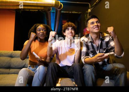 Trois jeunes amis victorieux heureux multiraciaux assis sur un canapé avec le premier geste de célébration fermé engagés dans une session de jeu vidéo amusante Banque D'Images