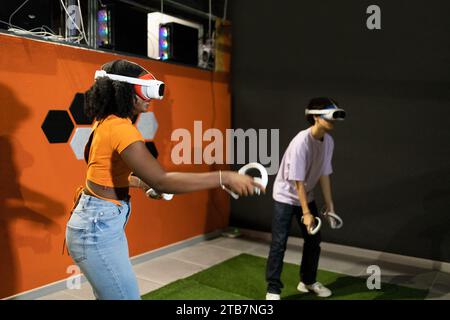 Vue latérale de jeunes amis multiraciaux engagés dans des jeux de réalité virtuelle portant des casques VR et utilisant des manettes portables dans un cadre intérieur Banque D'Images