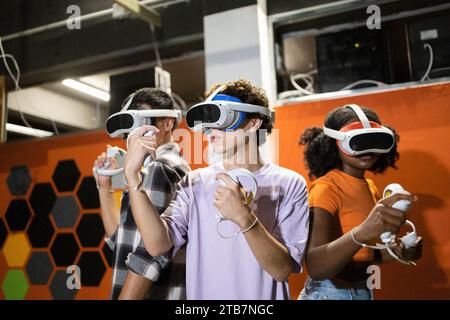 Jeunes amis multiraciaux entièrement immergés dans une session de jeu en réalité virtuelle brandissant des manettes et portant des casques VR Banque D'Images