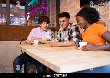 Joyeux groupe multiethnique d'amis avec des tasses de café dans un café confortable, mettant en valeur la navigation sur téléphone mobile Banque D'Images