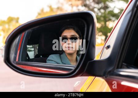 Réflexion de confiante jeune femme d'affaires caucasienne sérieuse portant des lunettes de soleil dans le miroir d'aile du véhicule moderne tout en se rendant au bureau Banque D'Images