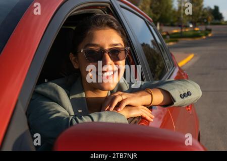 Portrait d'heureux attrayant caucasien jeune femme entrepreneur portant des lunettes de soleil et blazer assis dans la voiture orange et regardant par la fenêtre Banque D'Images