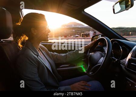 Portrait de jeune joyeux beau exécutif caucasien portant des lunettes de soleil et blazer conduisant une voiture moderne au coucher du soleil en ville Banque D'Images
