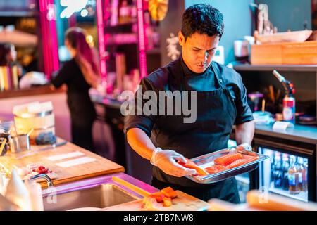 Cuisinier masculin confiant et concentré en uniforme noir et gants plaçant de la viande de saumon fraîchement coupée dans un plateau pour préparer le repas au bar à sushi illuminé Banque D'Images