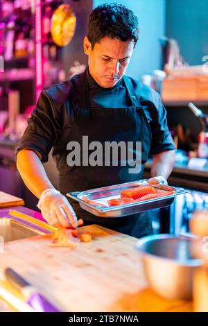 Cuisinier masculin confiant et concentré en uniforme noir et gants plaçant de la viande de saumon fraîchement coupée dans un plateau pour préparer le repas au bar à sushi illuminé Banque D'Images
