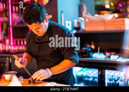 Chef masculin concentré en uniforme noir et gants utilisant des baguettes pour faire des rouleaux de sushi frais tout en travaillant dans la cuisine dans la boîte de nuit moderne illuminée Banque D'Images