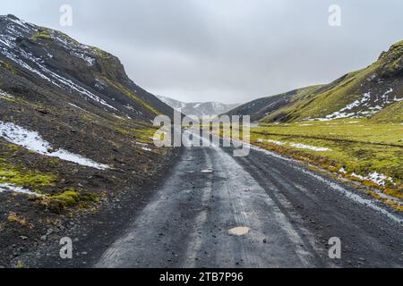 Une route de gravier désolée serpente à travers le paysage accidenté des hautes terres islandaises, flanquée de mousse verte, de falaises acérées et de restes de neige Banque D'Images