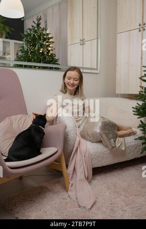 Une femme souriante dans une robe chic se loge sur un fauteuil rose avec son chat noir à côté d'elle dans un salon confortable et bien décoré. Banque D'Images