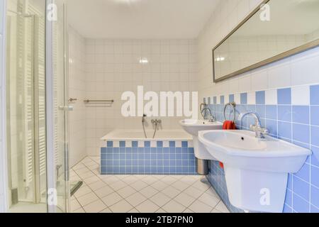 Long miroir monté au-dessus de deux lavabos près d'une baignoire dans une salle de bains spacieuse carrelée de bleu et blanc Banque D'Images