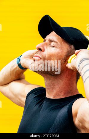 Vue latérale rapprochée de l'athlète masculin adulte moyen serein dans la casquette noire et les vêtements de sport avec les yeux fermés écoutant de la musique à travers des écouteurs contre le dos jaune Banque D'Images