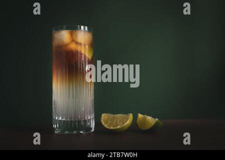 Cocktail highball sombre et orageux avec de la glace et des quartiers de citron vert sur une table en bois devant un fond vert foncé Banque D'Images