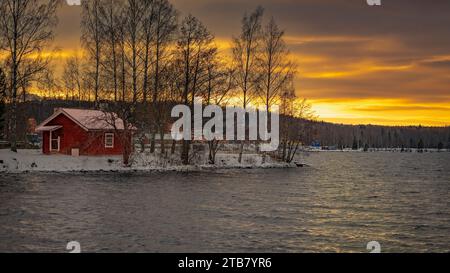 Une maison rouge vibrante se dresse seule sur la surface sereine d'un lac tranquille, entourée d'un paysage hivernal enneigé Banque D'Images