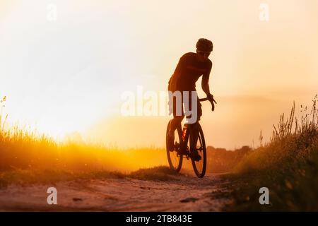 Silhouette jeune de cycliste sur un vélo de gravier roulant sur un sentier de poussière au coucher du soleil. Banque D'Images