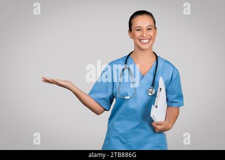 Médecin féminin européen excité tenant le presse-papiers montrant quelque chose sur la paume vide, produit publicitaire ou clinique Banque D'Images