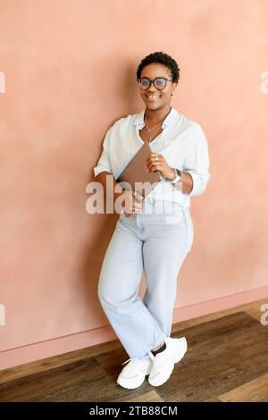 Portrait vertical pleine longueur d'ambitieuse femme chef de la direction entrepreneure se tient dans l'espace de bureau contemporain avec ordinateur portable, femme d'affaires souriante regardant la caméra, freelance dans le co-working sur le mur Banque D'Images