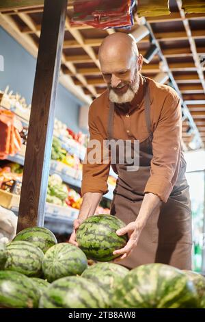 joyeux vendeur mature barbu souriant joyeusement et cueillant de la pastèque fraîche à l'épicerie Banque D'Images