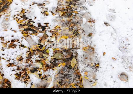 Neige précoce à l'automne. Feuilles jaunes tombées et chaussures imprimées sur la première neige mouillée. Route enneigée d'automne s'éloignant dans la distance. Concept de transition. Horizo Banque D'Images