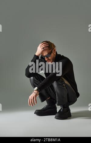 homme barbu dans des lunettes de soleil élégantes, pull col roulé et pantalon en cuir assis sur des hanches sur gris Banque D'Images