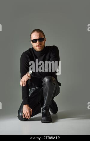 homme barbu dans des lunettes de soleil à la mode, pull col roulé et pantalon en cuir assis sur fond gris Banque D'Images