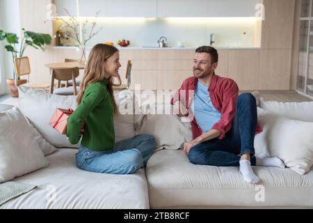 Heureuse petite amie souriante assise sur un canapé cachant derrière, félicitant joyeux petit ami heureux Banque D'Images