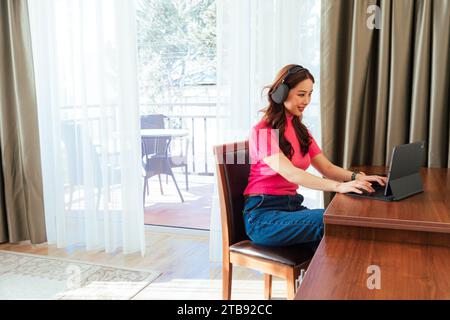 Femme asiatique indépendante souriante travaillant à la maison portant des écouteurs et en utilisant un ordinateur tablette et assis près de la table en bois Banque D'Images