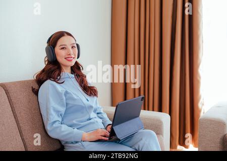 Gros plan de femme asiatique indépendante souriante travaillant à la maison portant des écouteurs et utilisant un ordinateur tablette et regardant la caméra Banque D'Images