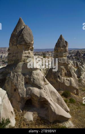 Grottes Maisons creusées dans les formations rocheuses volcaniques appelées cheminées de fées, contre un ciel bleu vif près de la ville de Goreme en arrière-plan ... Banque D'Images