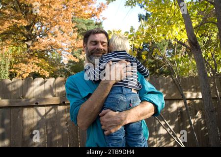 Homme tenant et embrassant son fils de trois ans dans leur cour à l'automne ; Lincoln, Nebraska, États-Unis d'Amérique Banque D'Images