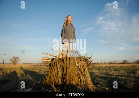 Fille debout sur une grande souche d'arbre dans la lumière du soleil dans la campagne regardant vers le bas la caméra ; Kearney, Nebraska, États-Unis d'Amérique Banque D'Images