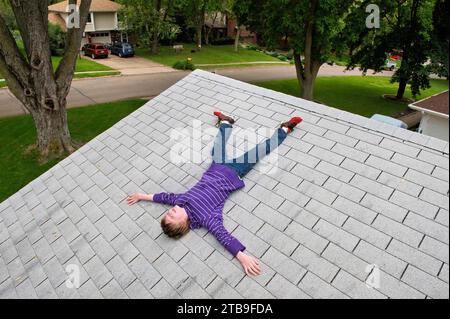 Garçon d'âge élémentaire allongé sur le toit en bardeaux d'une maison ; Elkhorn, Nebraska, États-Unis d'Amérique Banque D'Images