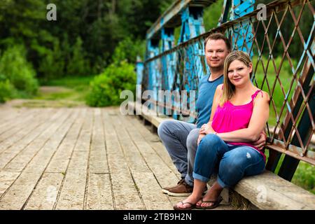 Portrait d'un couple assis ensemble sur un pont à chevalets en bois et regardant la caméra lors d'une promenade dans la nature dans un parc ; Edmonton, Alberta, Canada Banque D'Images